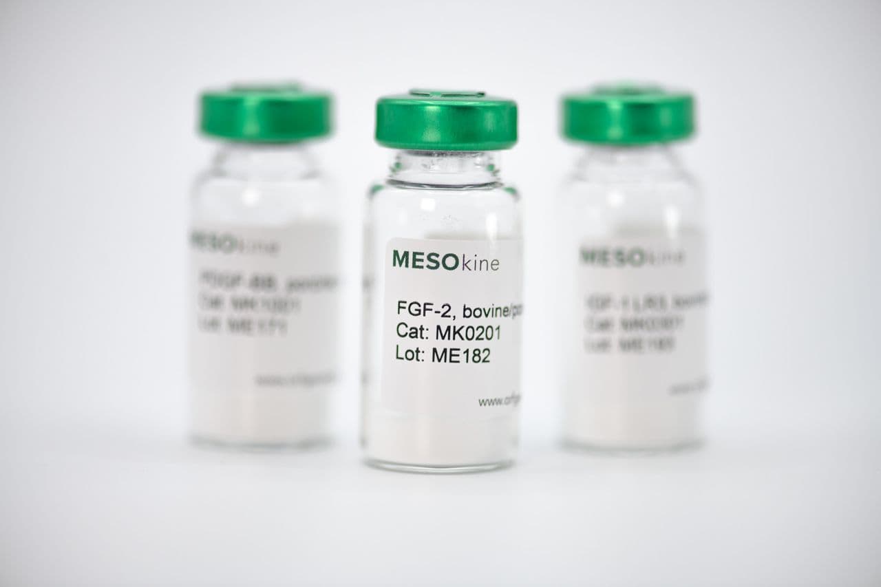 Three glass vials with green caps containing white powder, with the center vial labeled MESOkine FGF-2